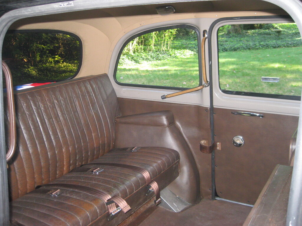 The rear passenger bench in worn brown leather, with the wide rear door open to a lawn and the grab handle above it.