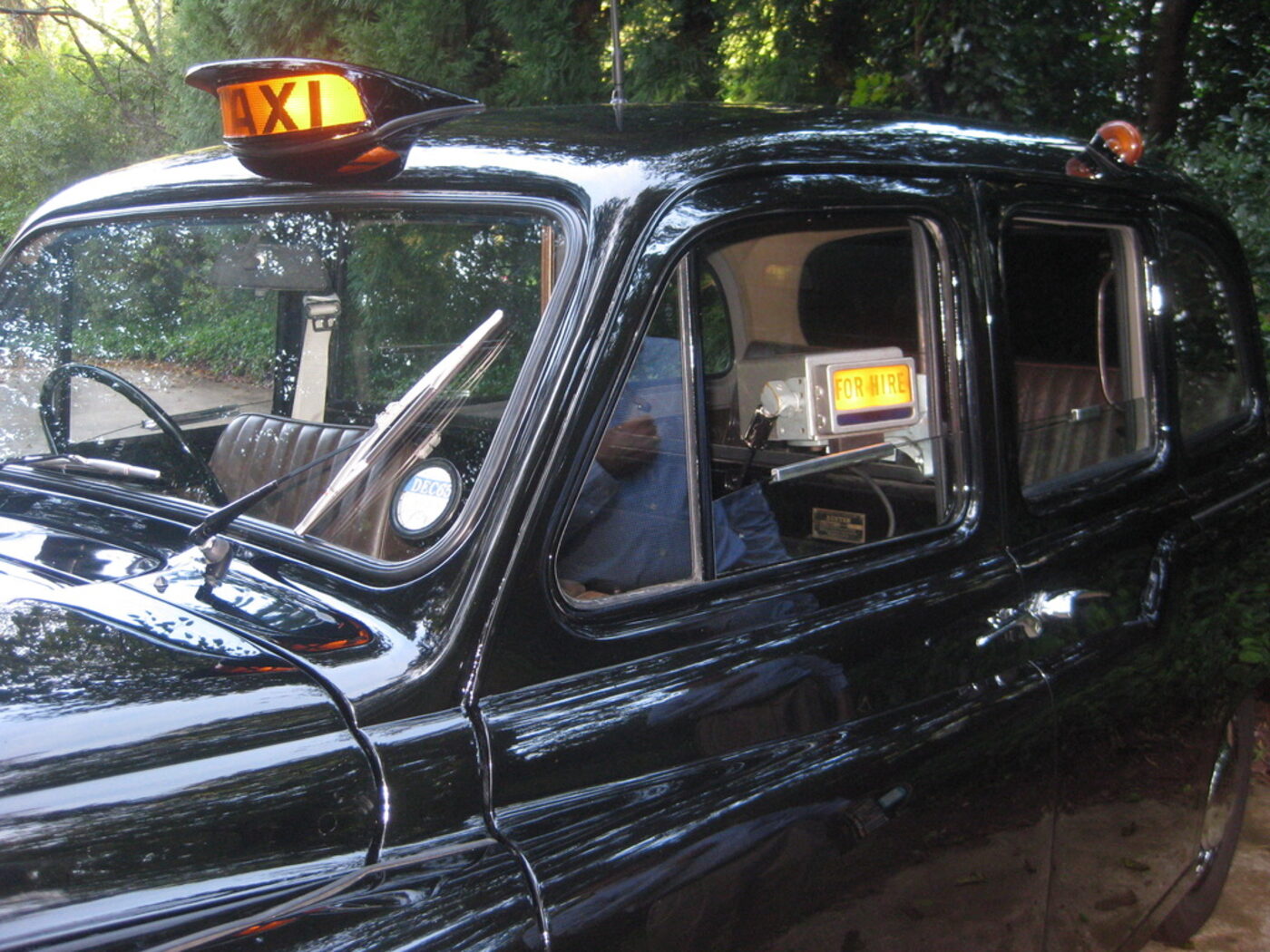 The side of the taxi showing the illuminated amber TAXI roof sign and the FOR HIRE panel glowing on the meter inside the cab.