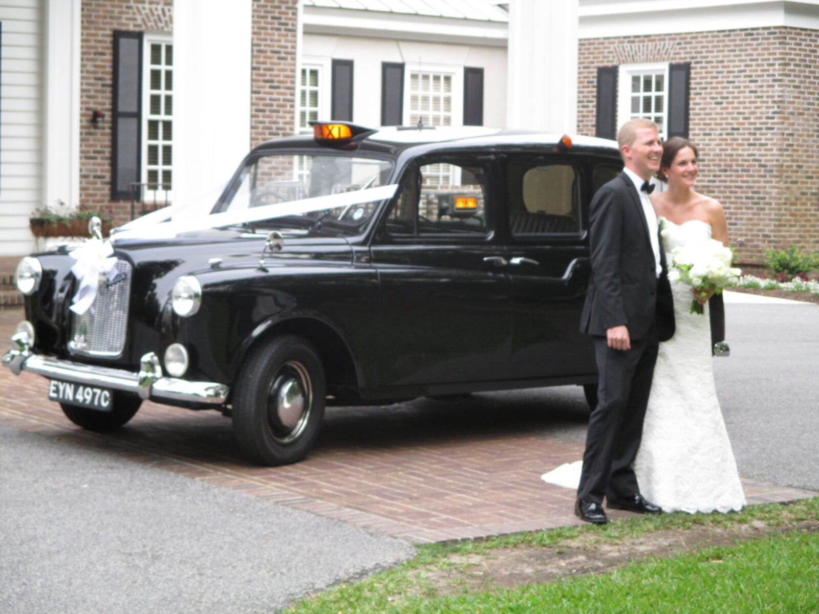 The black 1965 Austin FX4 taxi dressed with white ribbons across the bonnet, parked on a brick drive with a bride and groom standing beside it.